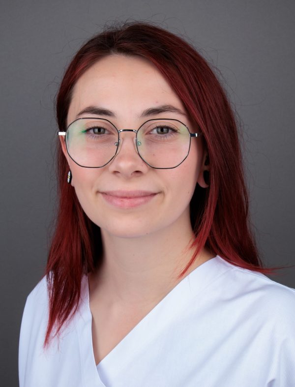 Portret žene s crvenom kosom i naočalama, obučena u bijelu majicu, na sivoj pozadini.