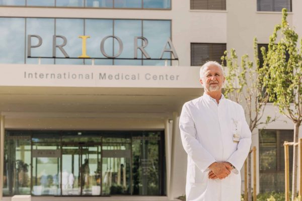 A gray-haired doctor in a white coat stands outside the Priora International Medical Center entrance, with the hospital’s signage visible behind him.