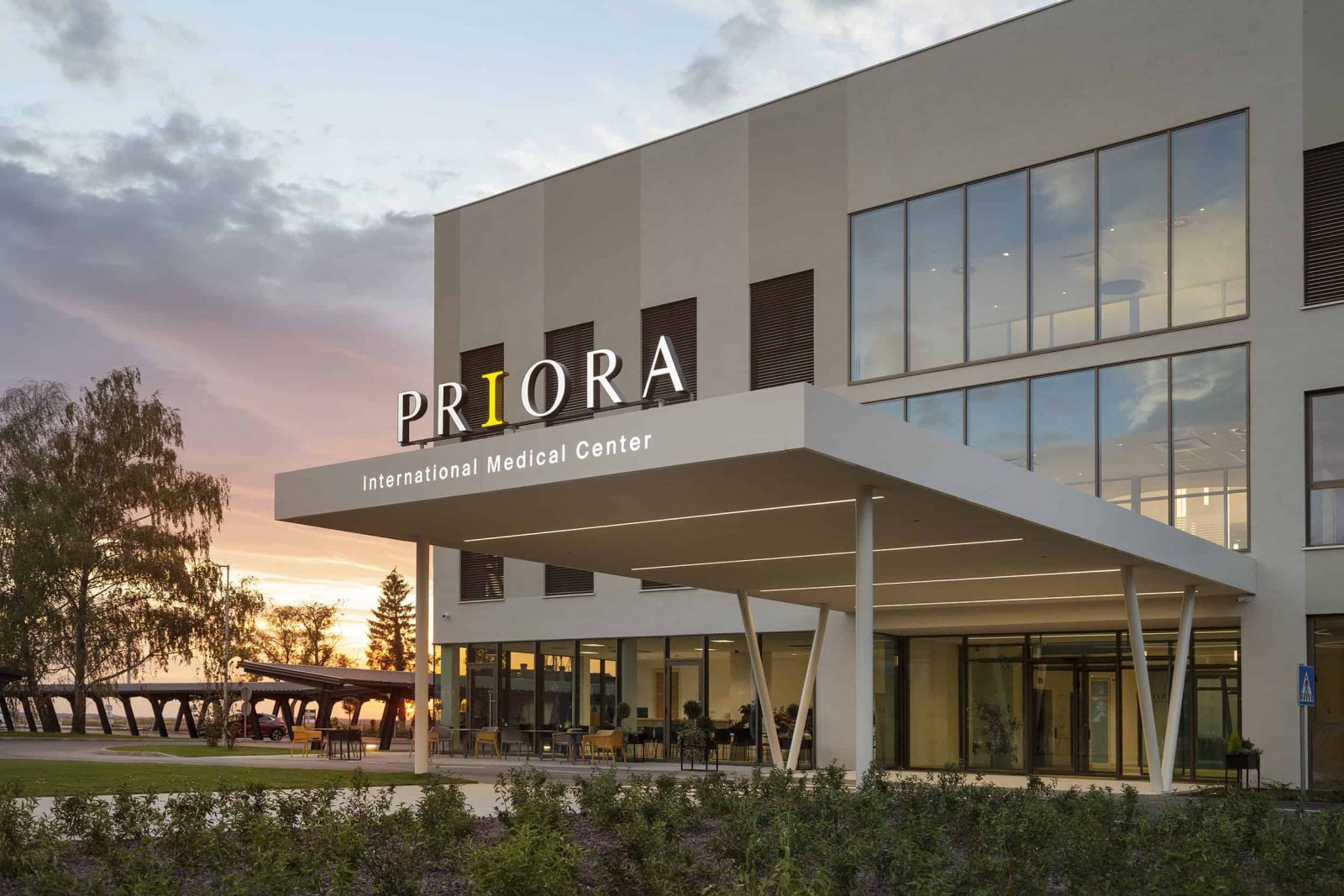 Exterior view of Priora International Medical Center during sunset, featuring large windows and a modern entrance with a canopy.