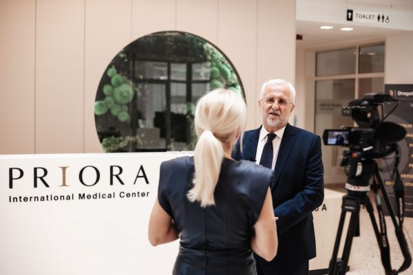 Osoba sa sijedom kosom i bradom daje intervju ispred kamere u medicinskom centru Priora, dok mu sugovornica stoji leđima okrenuta kameri.