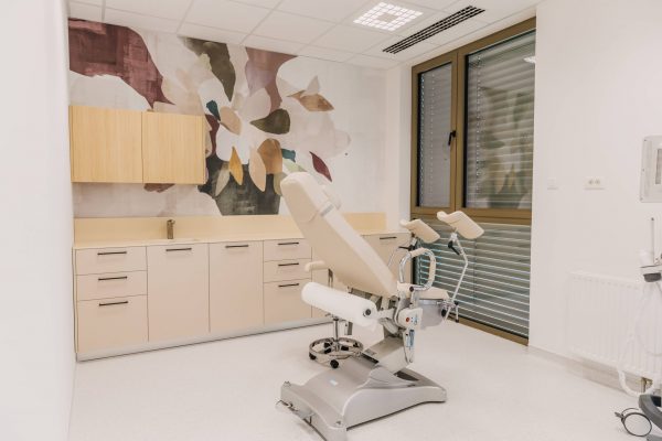 Moderna ginekološka ambulanta s ginekološkom stolicom, bež ormarićima i zidnim muralom s cvjetnim motivom. Prostorija ima veliki prozor sa žaluzinama i svijetao, čist dizajn.