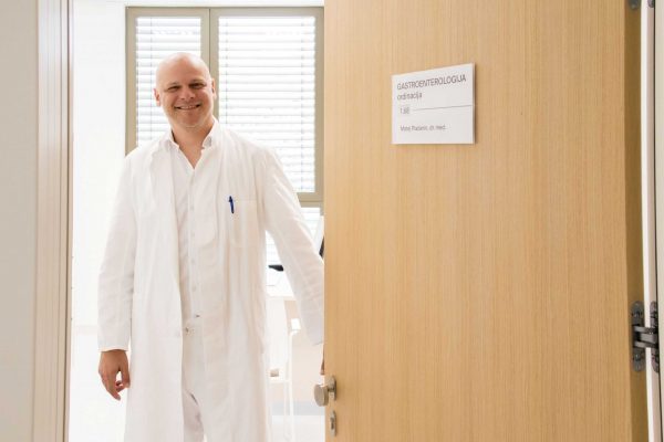 Liječnik u bijeloj kutiji stoji nasmijano na vratima ordinacije gastroenterologije s natpisom Matej Plazanić, dr. med.
