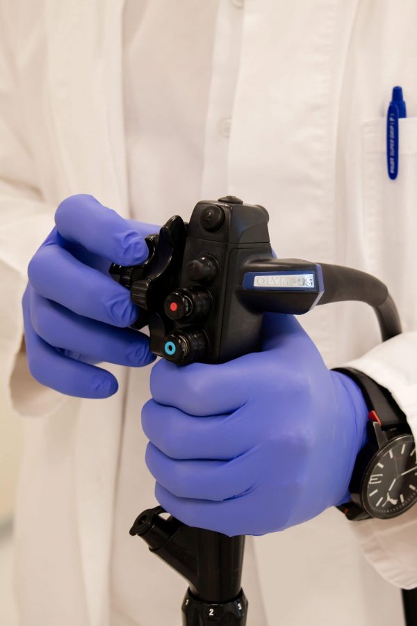 Osoba u bijelom medicinskom ogrtaču i plavim rukavicama drži endoskop.