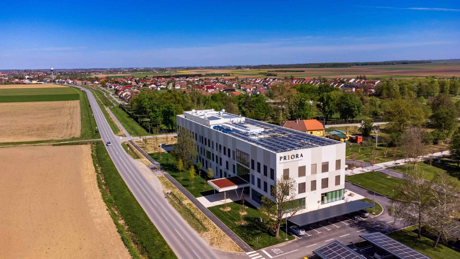 Zgrada s natpisom 'PRIORA' okružena zelenilom i cestom, smještena uz obradivo polje i naselje.