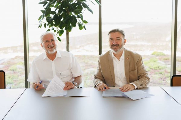 Dva muškarca sjede za stolom s dokumentima i kemijskom olovkom, nasmiješeni, u svijetloj prostoriji s biljkama u pozadini.