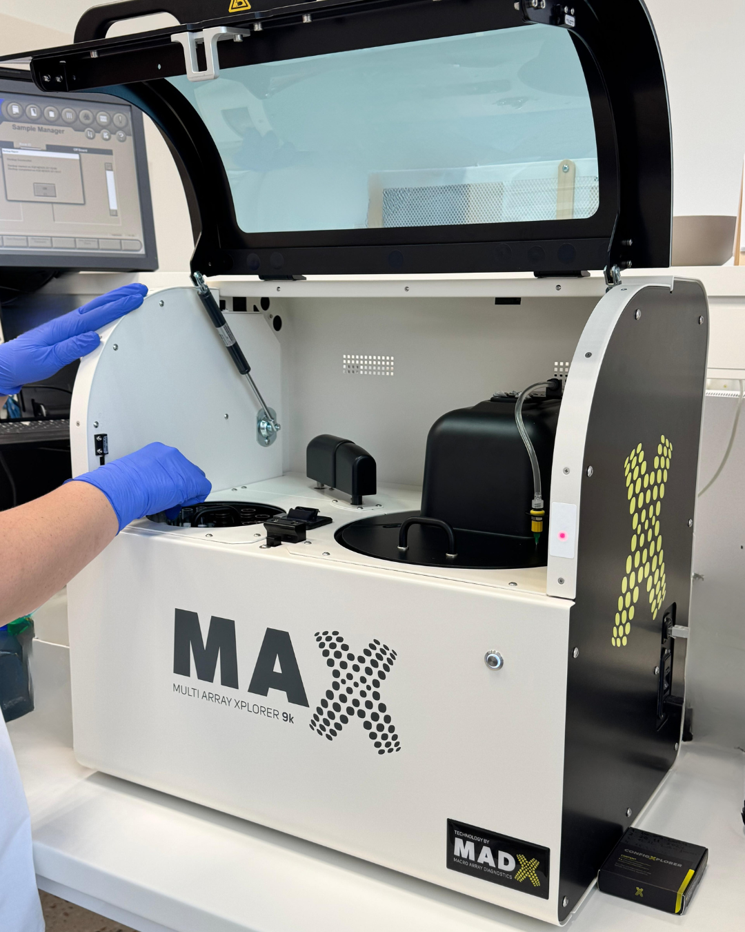 Osoba u zaštitnim rukavicama rukuje bijelim laboratorijskim uređajem s otklopljenim poklopcem, označenim natpisom 'MAX Multi Array Xplorer 9k'.