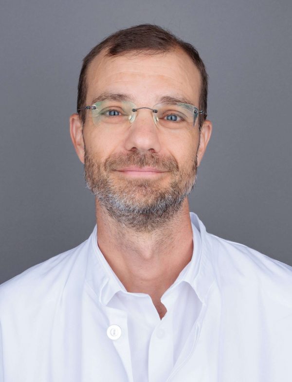 Muškarac s naočalama u bijelom laboratorijskom ogrtaču, s blagim osmijehom, na sivoplavoj pozadini.