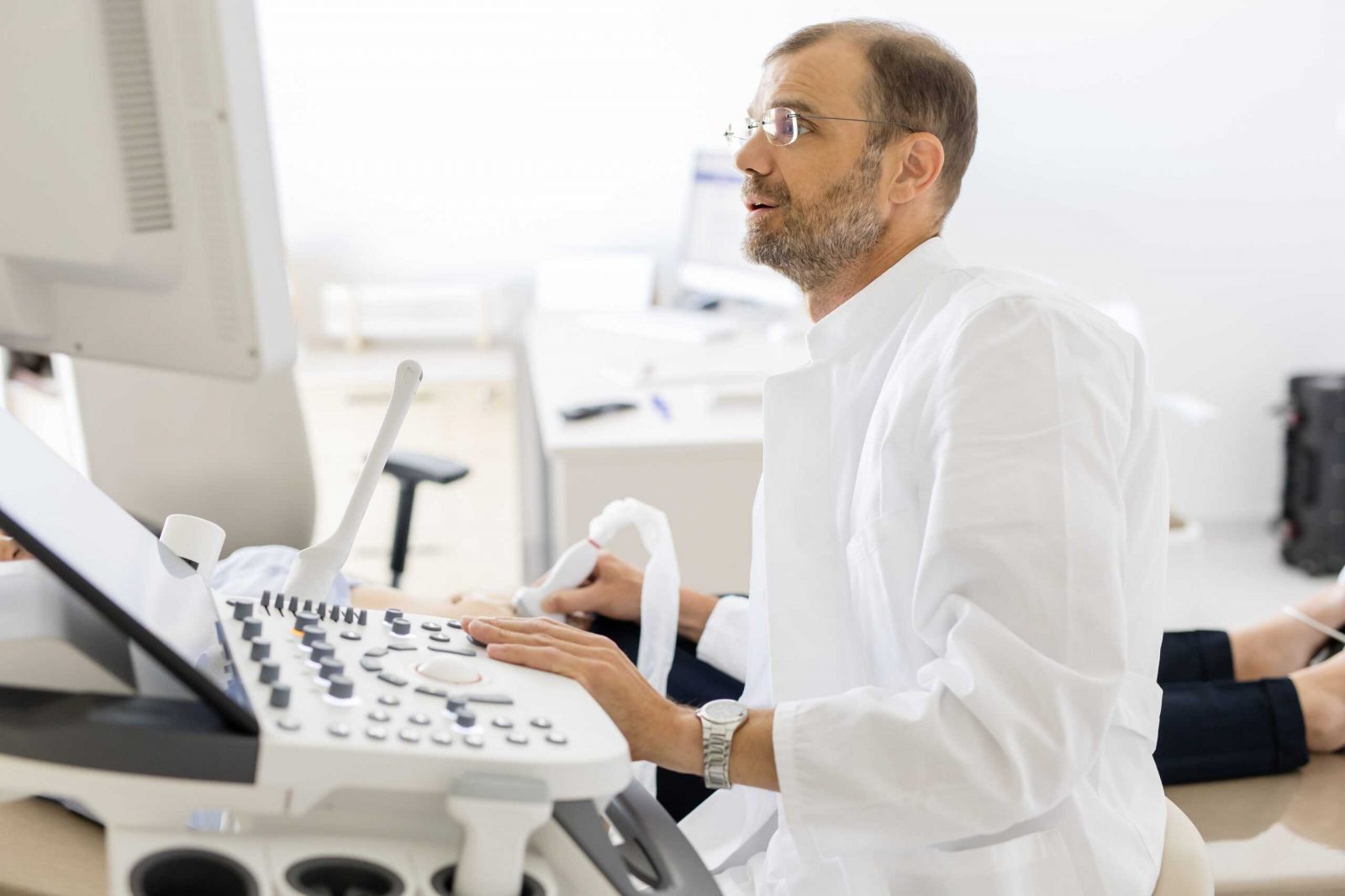 Muškarac u bijelom medicinskom ogrtaču izvodi ultrazvučni pregled koristeći aparat u svijetloj ordinaciji.