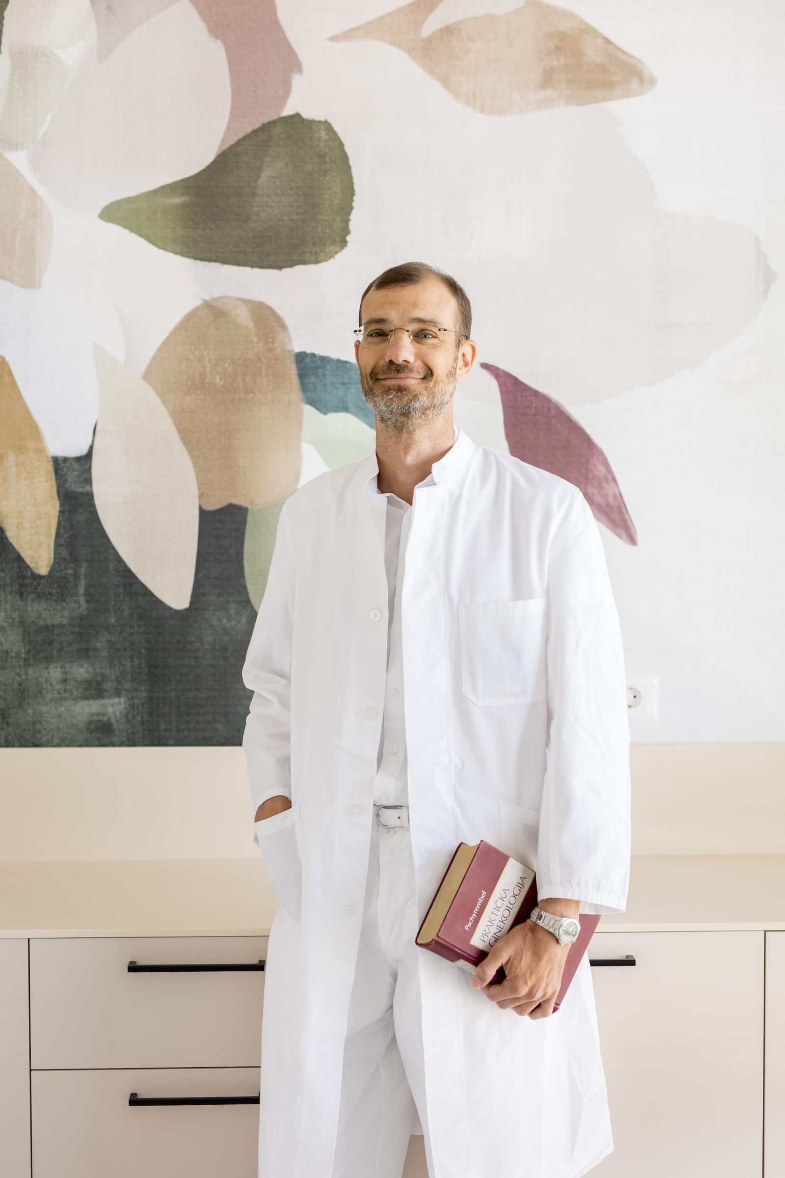 Muškarac u bijeloj kutama, s knjigom u ruci, stoji uz šareni zidni uzorak.