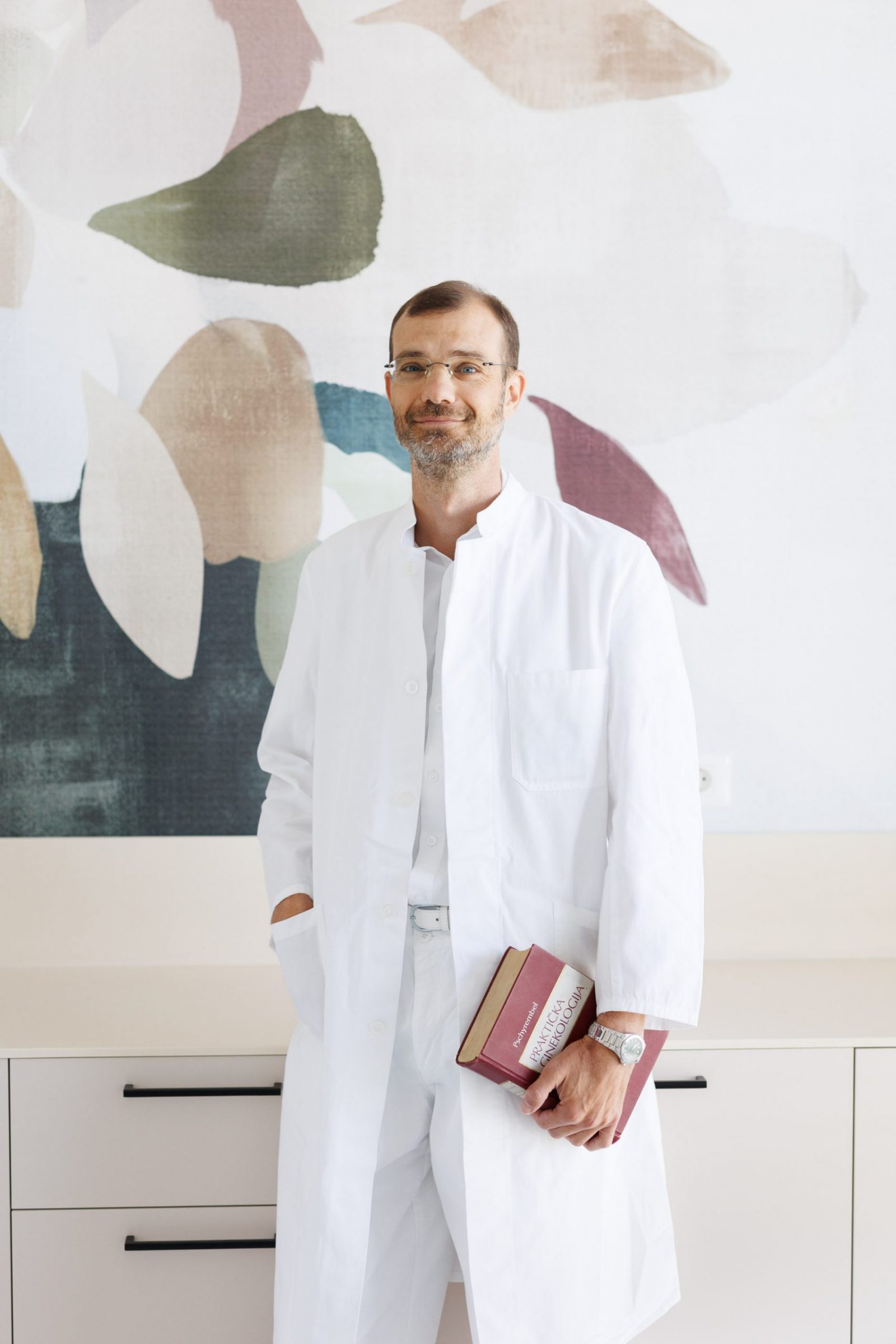 Smiling doctor in a white lab coat holding a red medical textbook, standing in a bright clinic with abstract wall art.