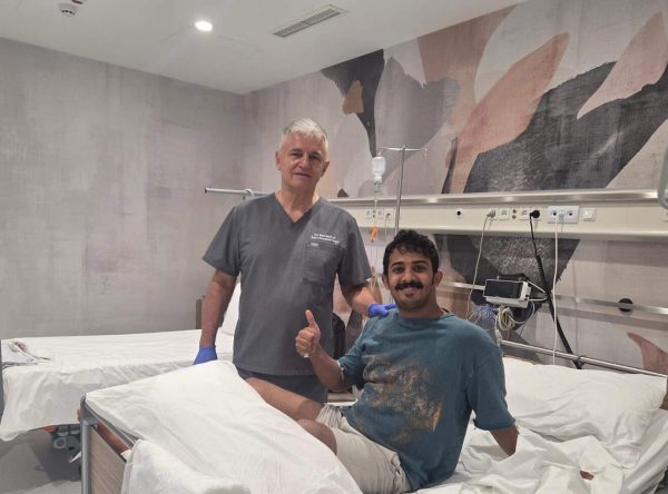 Osoba u bolničkom krevetu daje znak odobravanja palcem gore, dok pored nje stoji medicinski radnik u sivoj uniformi; prostorija je moderno uređena s apstraktnim motivima na zidu.