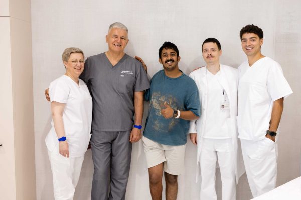 Skupina od pet osoba stoji zajedno, od kojih su četiri u medicinskoj odjeći, dok jedan nosi ležernu odjeću i pokazuje znak odobravanja palcem gore.