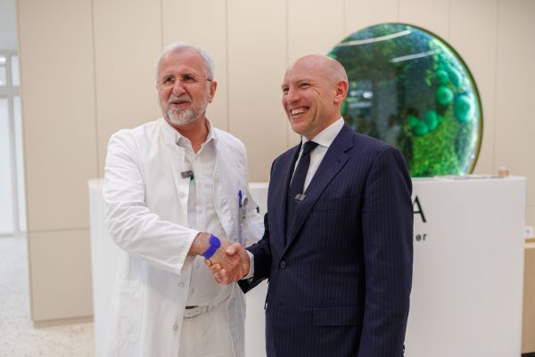 Dvojica muškaraca se rukuju; jedan nosi bijeli laboratorijski ogrtač, a drugi tamno odijelo. Iza njih je okrugla, ukrasna instalacija u zelenim nijansama.