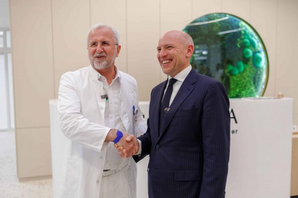 A smiling doctor in a white lab coat shakes hands with a man in a dark pinstripe suit in a modern lobby, both wearing clip-on microphones, with a large circular green art piece on the wall behind them.