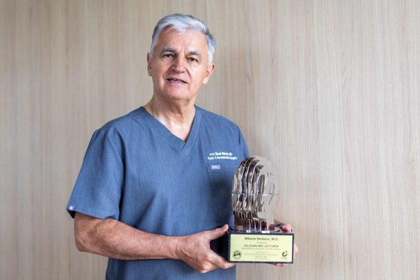 Stariji muškarac s sijedom kosom, u plavom medicinskom odijelu, drži trofej oblikovan kao stilizirana glava, stoji ispred svijetlosmeđe drvene pozadine.