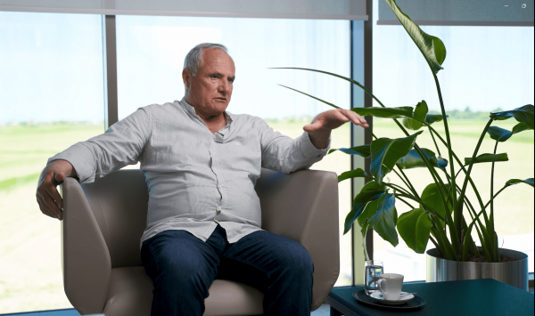 Stariji muškarac sjedi u fotelji i razgovara, s velikom biljkom sa zelenim lišćem pored sebe i šalicom kave na stolu.