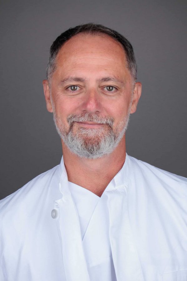 Muškarac s kratkom sivom kosom i bradom u bijeloj liječničkoj uniformi, smiješi se na sivoj pozadini.