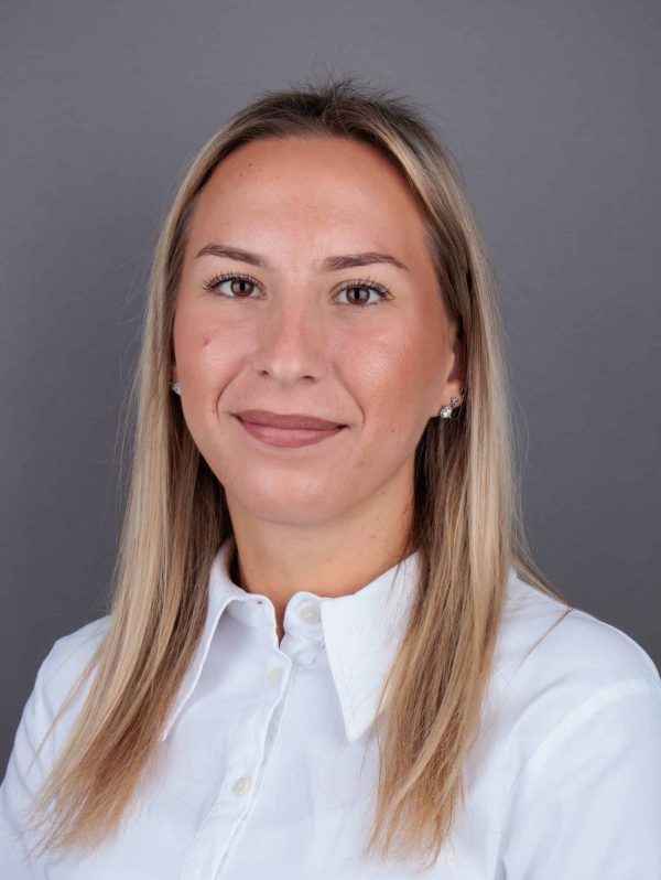 Žena s dugom plavom kosom i bijelom košuljom, pozira ispred sive pozadine.