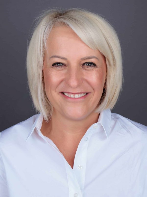 Žena svijetle kose i bijele košulje, nasmiješena, siva pozadina.