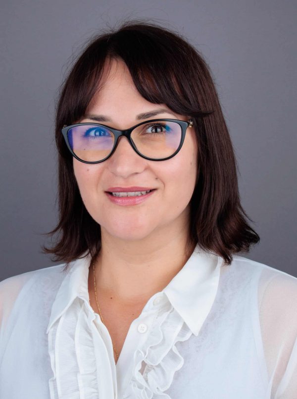 Žena s kratkom tamnom kosom i naočalama, nosi bijelu bluzu s volanima, pozira ispred sive pozadine.