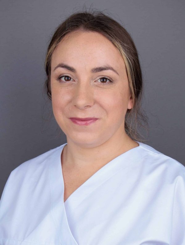 Žena svezane kose, odjevena u bijelu odjeću, smiješi se ispred sive pozadine.