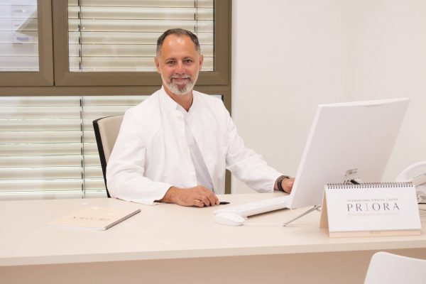 Muškarac u bijeloj medicinskoj kuti sjedi za stolom s računalom, nasmiješen, s posudom za dokumente na kojoj je natpis 'International Medical Center PRIORA'.