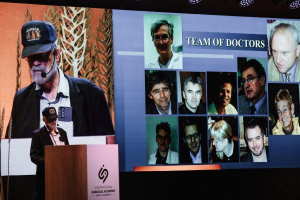Predavač s kapom i mikrofonom stoji za govornicom na konferenciji International Surgical Academy, dok se na velikom ekranu prikazuje slajd "TEAM OF DOCTORS" s nizom portreta liječnika.