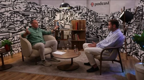 Dvojica muškaraca sjede u studiju podcasta i razgovaraju, na foteljama oko niskog stolića s čašama vode, uz policu s knjigama i nagradama te crno-bijeli grafički zid u pozadini.