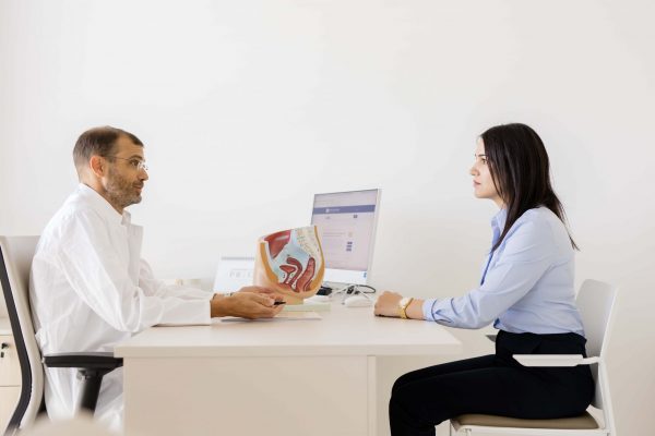 Liječnik u bijeloj kuti savjetuje ženu u ordinaciji, s anatomskim modelom ženske zdjelice i reproduktivnog sustava na stolu te računalnim monitorom u pozadini.