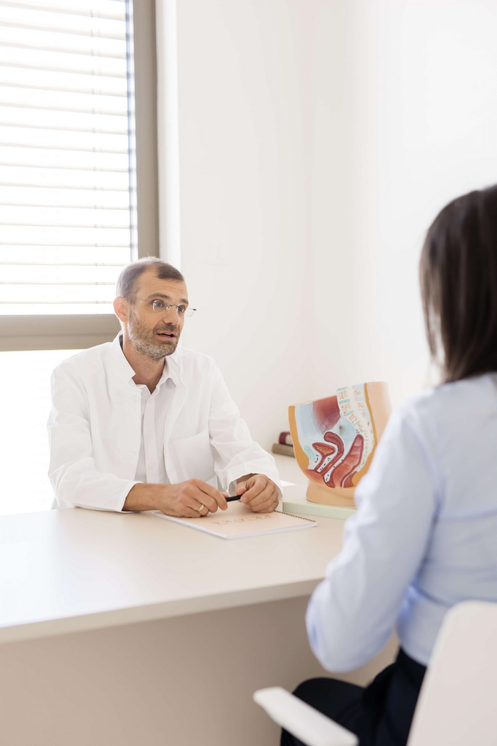 Liječnik u bijeloj kuti razgovara s pacijentom u ordinaciji, a na stolu je anatomski model zdjelice i rektuma.