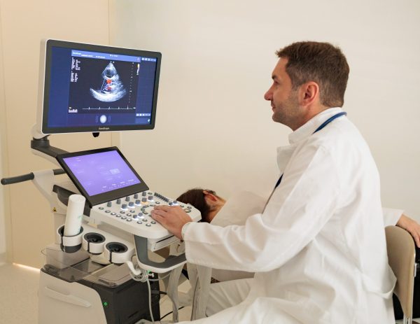 Liječnik u bijeloj kuti izvodi ultrazvuk srca pacijentu, dok se na monitoru vidi ehokardiogram s Doppler prikazom protoka.