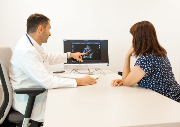 Liječnik u bijeloj kuti tijekom konzultacija pokazuje pacijentu na monitoru ehokardiogram s Doppler prikazom u svijetloj ordinaciji.