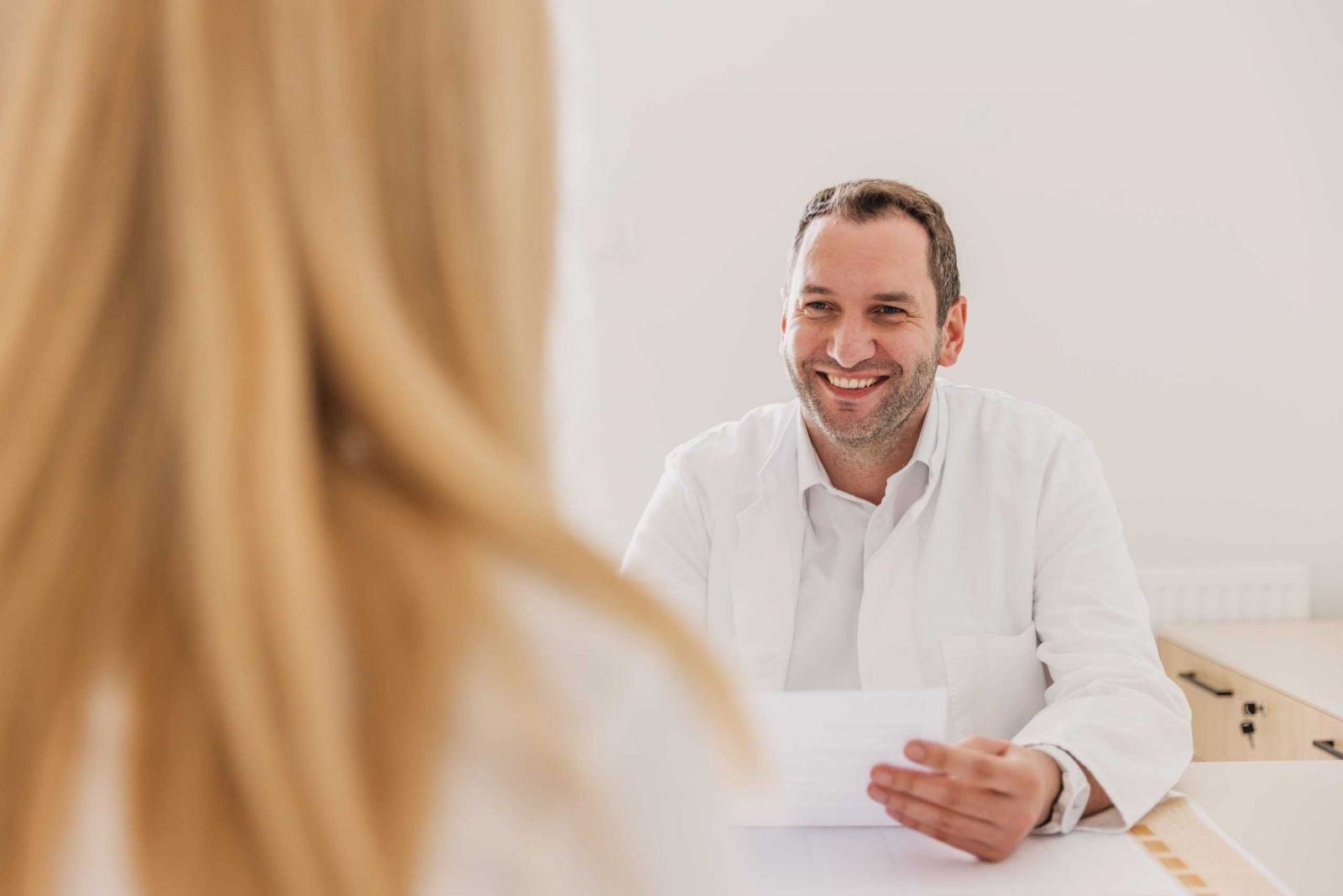 Smiling doctor in a white coat sits at a desk holding a paper during a consultation with a patient, whose blonde hair is visible in the foreground.