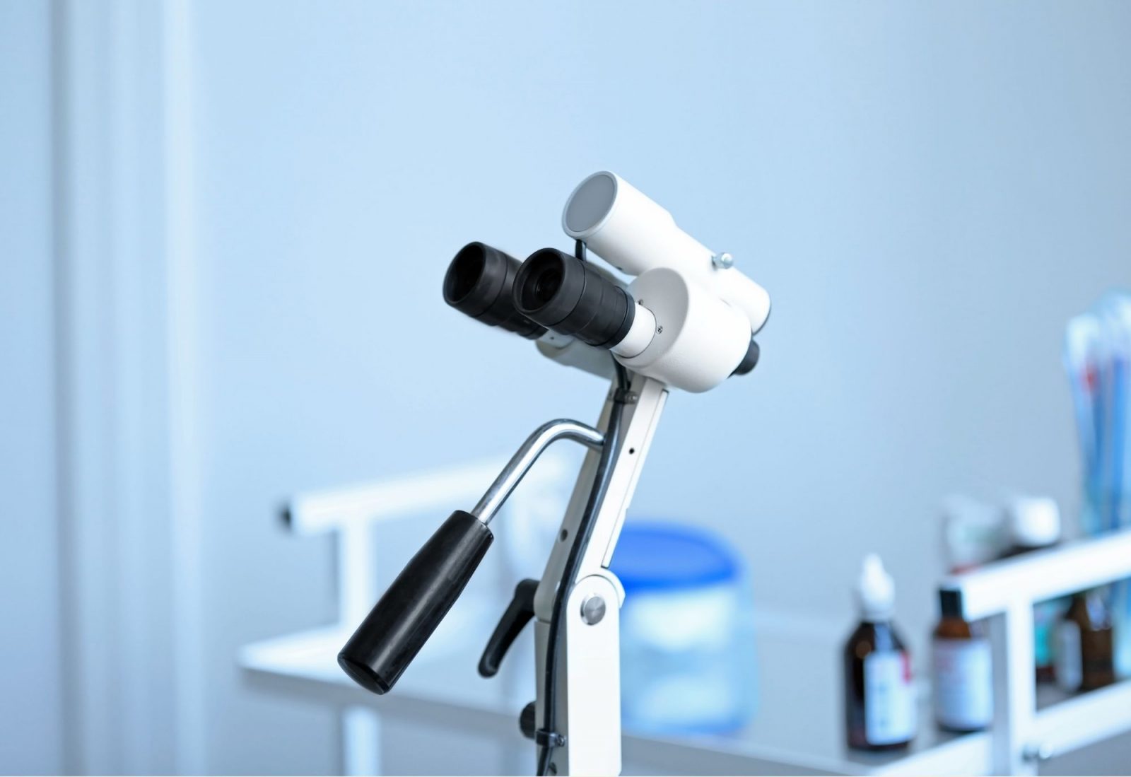 Kolposkop s dvostrukim okularom i ručkom u svijetloj ambulanti, s bočicama i medicinskim priborom zamućenima u pozadini.