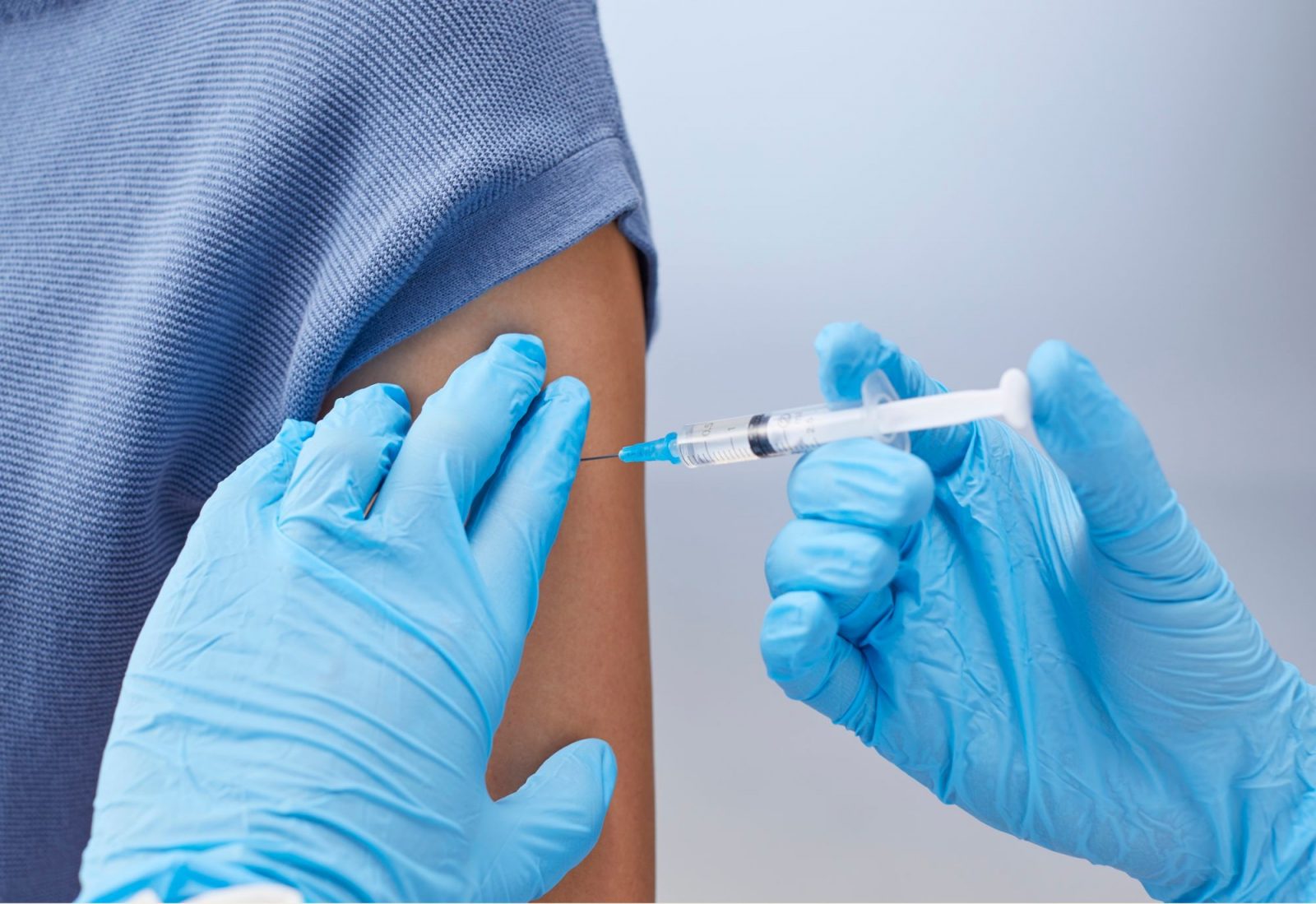 Gloved healthcare worker administers a vaccine injection into a person’s upper arm with their sleeve rolled up.