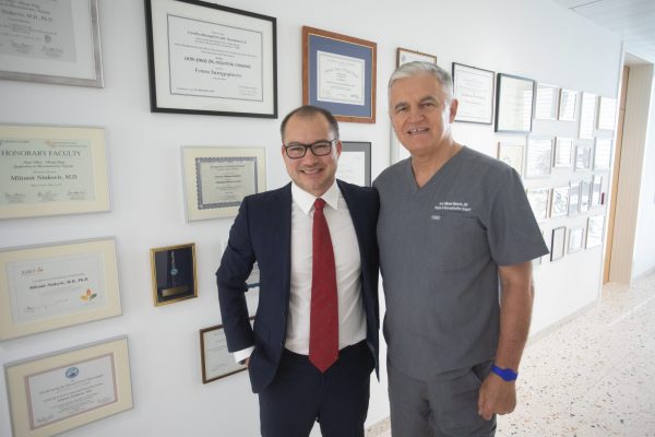 Two professionals stand side by side, smiling in a bright hallway lined with framed certificates and awards; one wears a navy suit with a red tie, the other gray medical scrubs.
