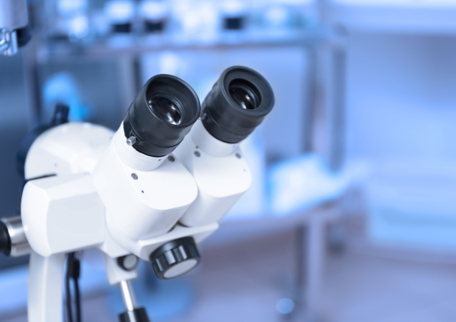 Krupan plan binokularnog mikroskopa s dva okulara i kotačićem za fokusiranje, sa zamućenom laboratorijskom pozadinom u hladnim plavim tonovima.