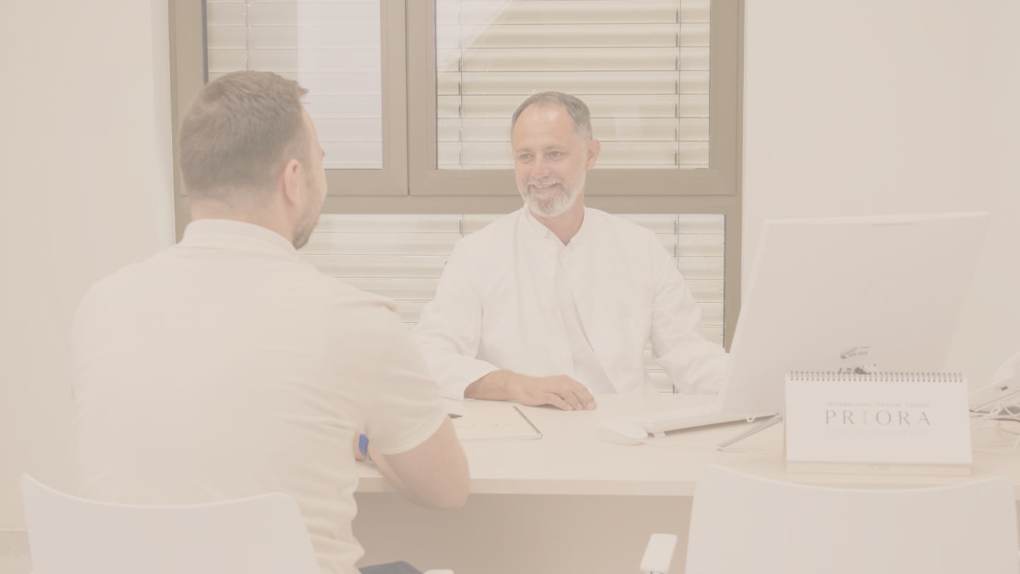 Liječnik u bijeloj kuti nasmiješeno razgovara s pacijentom preko stola u modernoj ordinaciji, s računalom i stolnim kalendarom „PRIORA“ u prvom planu.