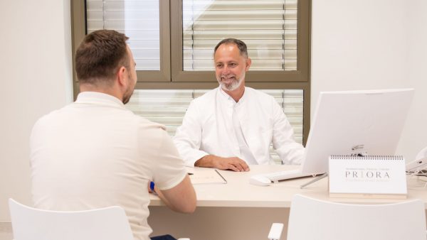 Liječnik u bijeloj kuti razgovara s pacijentom preko radnog stola u svijetloj ordinaciji, ispred računala i stonog kalendara s natpisom „PRIORA”.