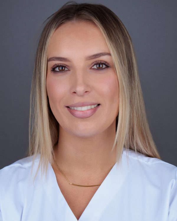 Portret nasmiješene osobe svijetle kose u bijeloj bluzi, s tankom zlatnom ogrlicom, na neutralnoj sivoj pozadini.