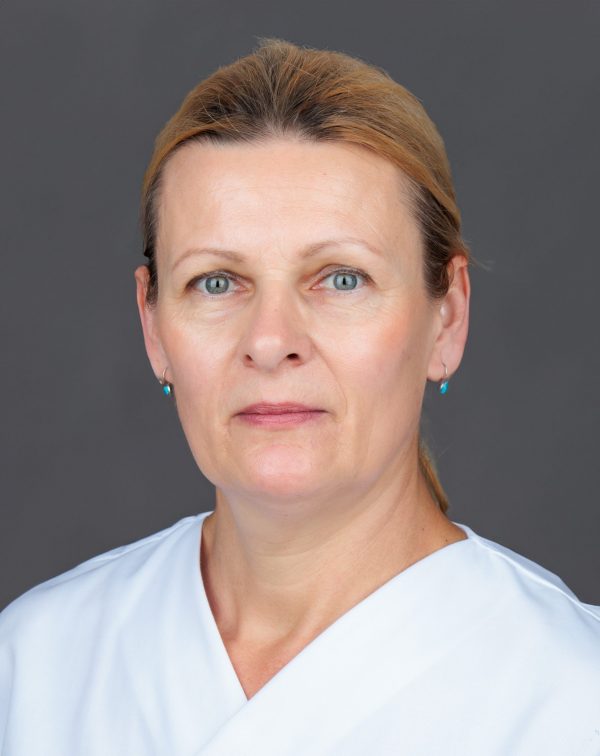 Bliski portret žene srednjih godina sa svijetlosmeđom podignutom kosom i svijetloplavim očima, u bijeloj bluzi s V-izrezom i sitnim tirkiznim naušnicama, neutralnog izraza, na sivoj pozadini.