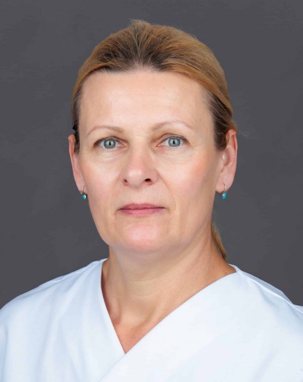 Bliski portret žene srednjih godina sa svijetlosmeđom podignutom kosom i svijetloplavim očima, u bijeloj bluzi s V-izrezom i sitnim tirkiznim naušnicama, neutralnog izraza, na sivoj pozadini.