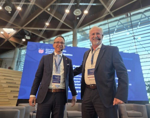 Dvojica nasmiješenih muškaraca u odijelima s headset mikrofonima stoje na konferencijskoj pozornici ispred velike plave pozadine s grbom Republike Hrvatske i tekstom ministarstva, noseći akreditacije oko vrata.