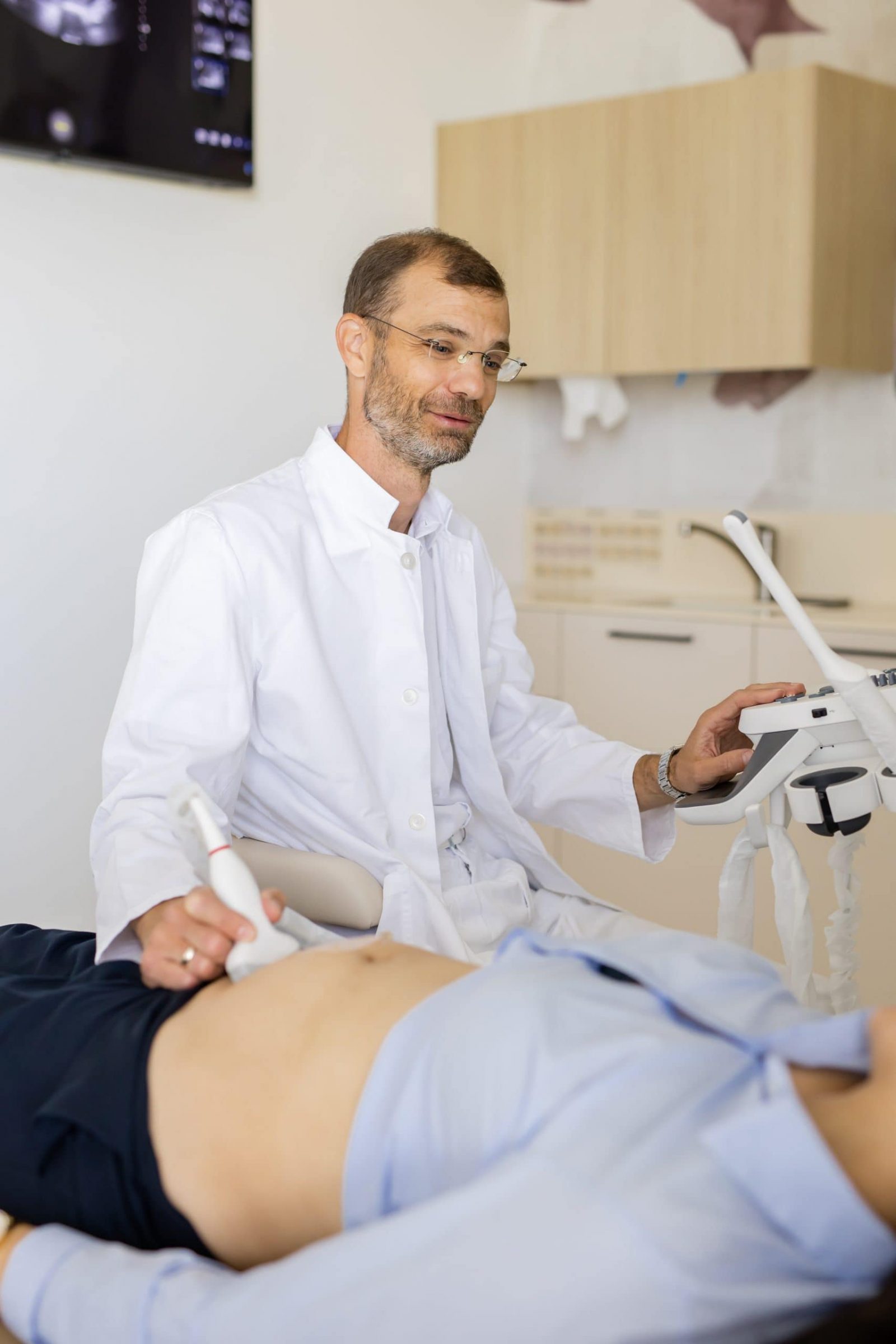 Liječnik u bijeloj kuti u ordinaciji izvodi ultrazvučni pregled trbuha pacijenta, držeći sondu na koži i upravljajući konzolom dok je ekran u pozadini.