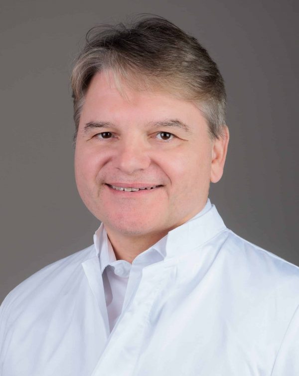 Nasmiješeni muškarac srednjih godina s kratkom prosijedom kosom u bijeloj kuti, portret na jednobojnoj sivoj pozadini.