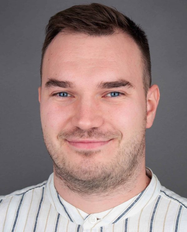 Portret muškarca s kratkom smeđom kosom, plavim očima i laganom bradicom, u bijeloj prugastoj košulji pred sivom pozadinom.