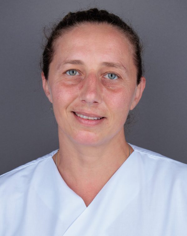 Portret žene u bijeloj medicinskoj uniformi s blagim osmijehom, plavim očima i kosom zavezanom unazad, na sivoj pozadini.