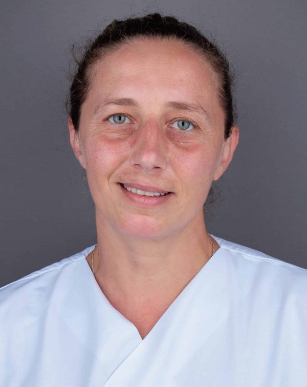 Portret žene u bijeloj medicinskoj uniformi s blagim osmijehom, plavim očima i kosom zavezanom unazad, na sivoj pozadini.