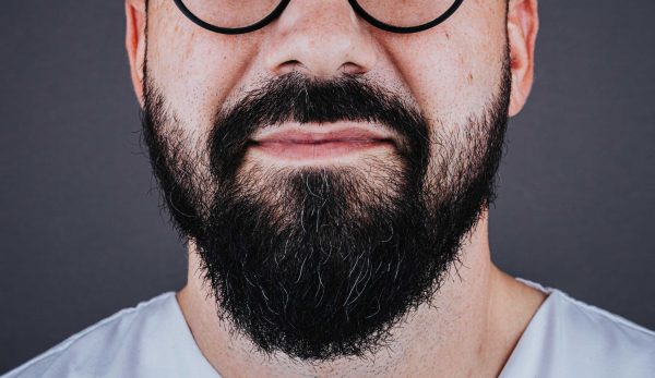Close-up of the lower face of a person with round glasses and a thick dark beard with a few gray strands, lips closed in a slight smile against a dark background.
