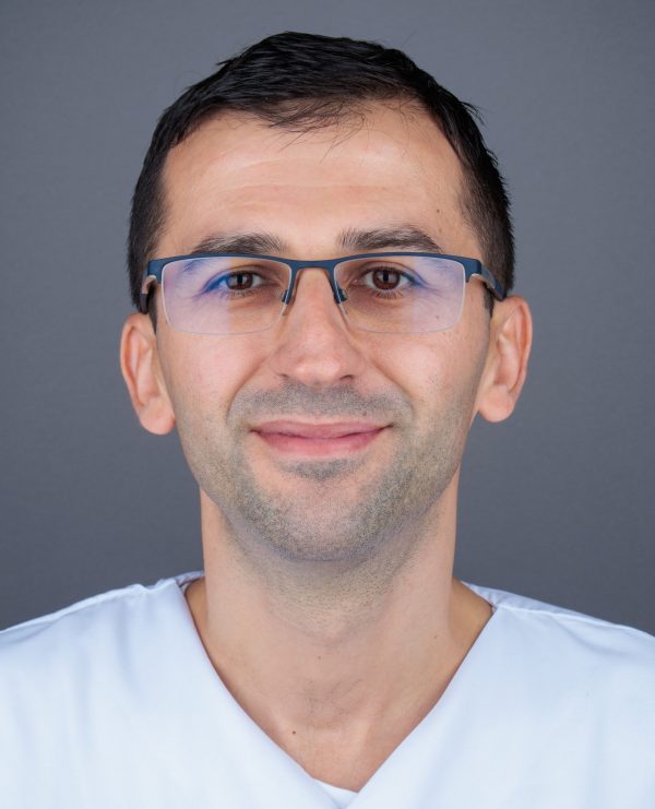 Portret muškarca s kratkom tamnom kosom i tankim plavkastim naočalama, u bijeloj majici nalik bolničkoj uniformi, s blagim osmijehom na sivoj pozadini.